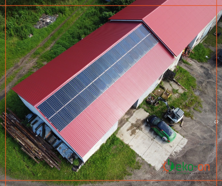 Widok z drona na budynek z czerwoną blachodachówką i panelami słonecznymi w Krakowie, obok zaparkowany zielony samochód i mała koparka.