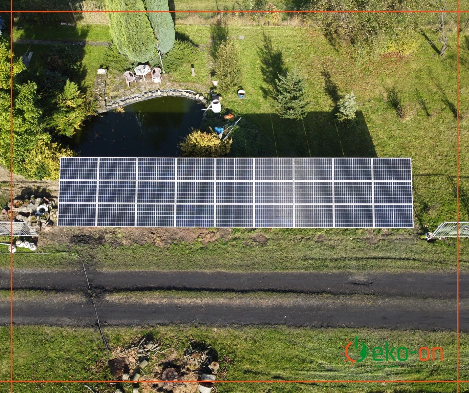 Widok z drona na panele słoneczne zainstalowane na trawniku obok ścieżki, w tle oczko wodne z miejscem do siedzenia.