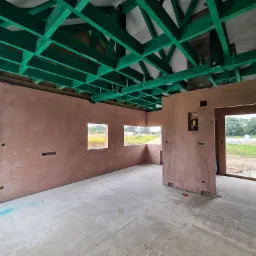 Wnętrze surowego domu w trakcie budowy: ściany otynkowane na różowo, zielona drewniana konstrukcja dachu, widoczne otwory okienne i drzwiowe, surowy beton na podłodze.