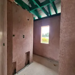 Wnętrze domu z keramzytu w trakcie budowy, widoczne ściany z surowego keramzytu z otworami na instalacje, zielona drewniana konstrukcja dachu i okno z widokiem na zieleń.