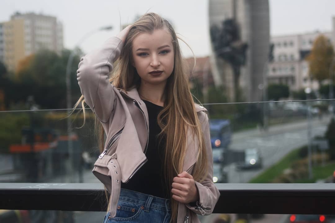 Portret młodej kobiety w beżowej kurtce i dżinsach, pozującej na tle miejskiego krajobrazu z budynkami i ruchem ulicznym, z ręką we włosach i ciemną szminką.