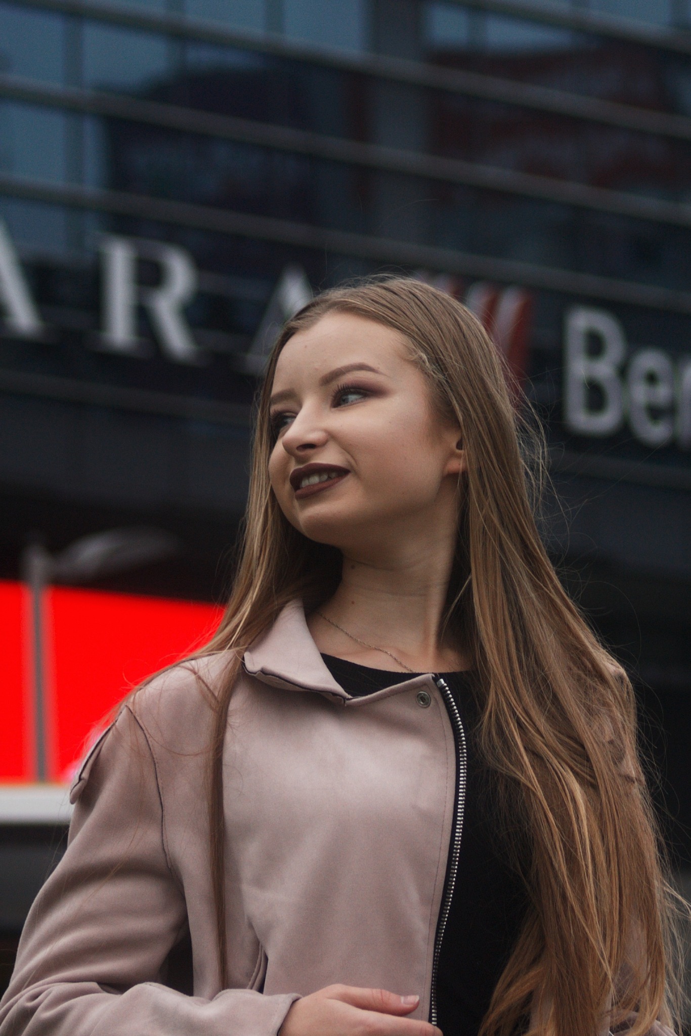 Portret młodej kobiety w jasnoróżowej kurtce na tle budynku z napisem, spoglądającej w bok.