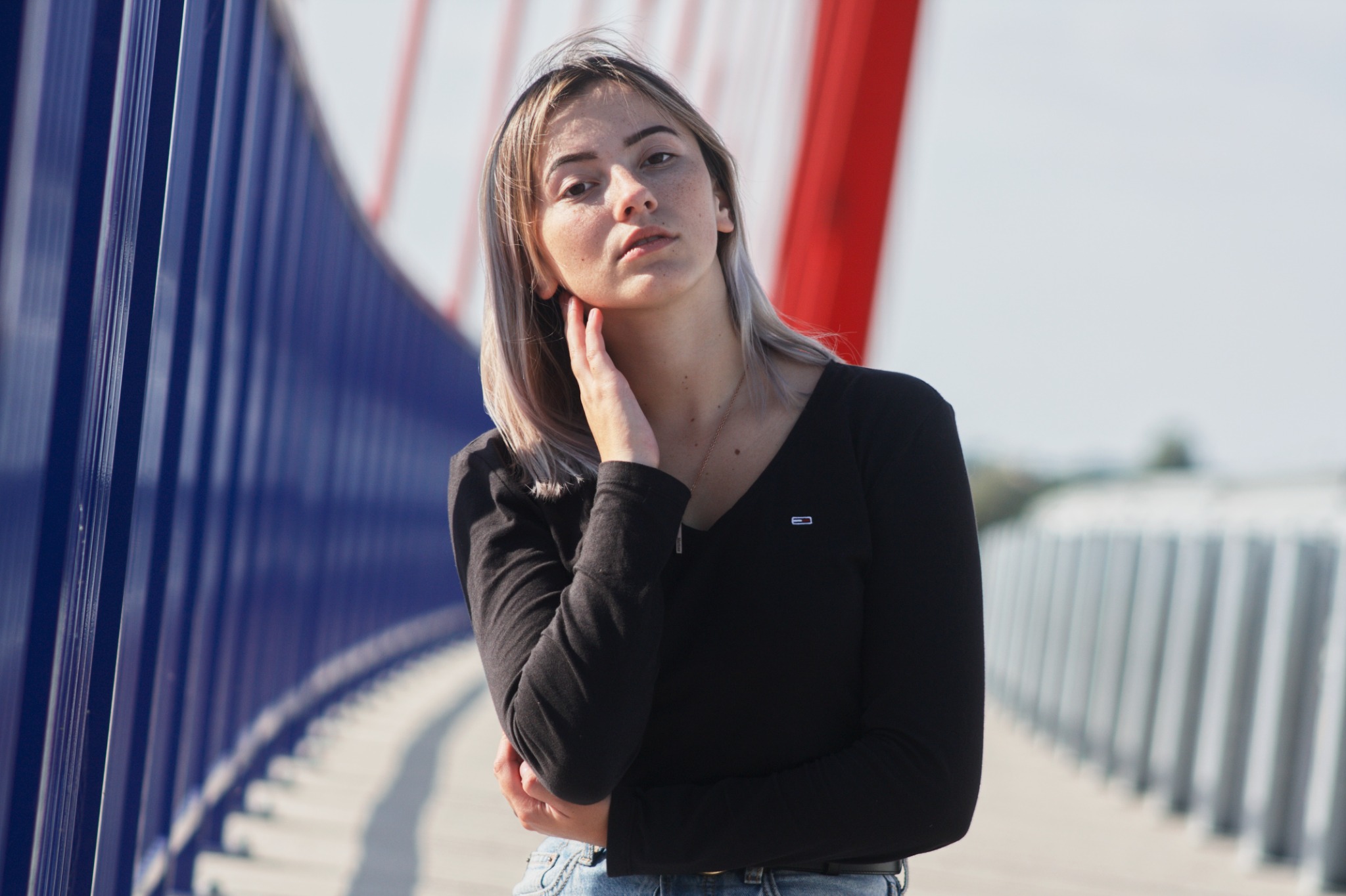 Portret kobiety o jasnych włosach i piegach, pozującej na tle mostu z niebieskimi i czerwonymi elementami konstrukcyjnymi.