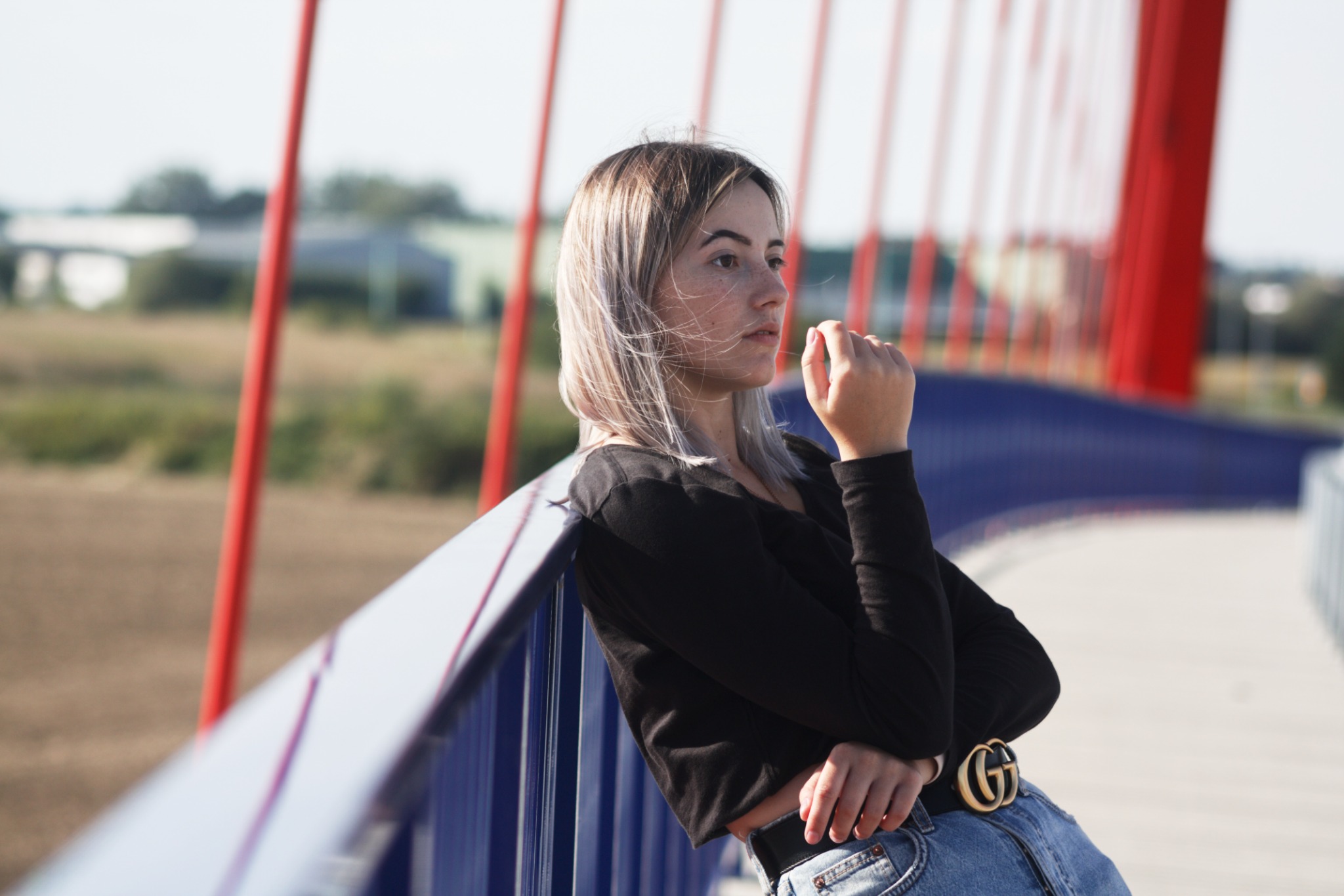 Młoda kobieta z piegami, oparta o niebieską barierkę mostu z czerwonymi elementami, ubrana w czarną bluzkę i dżinsy z paskiem Gucci, patrzy w dal