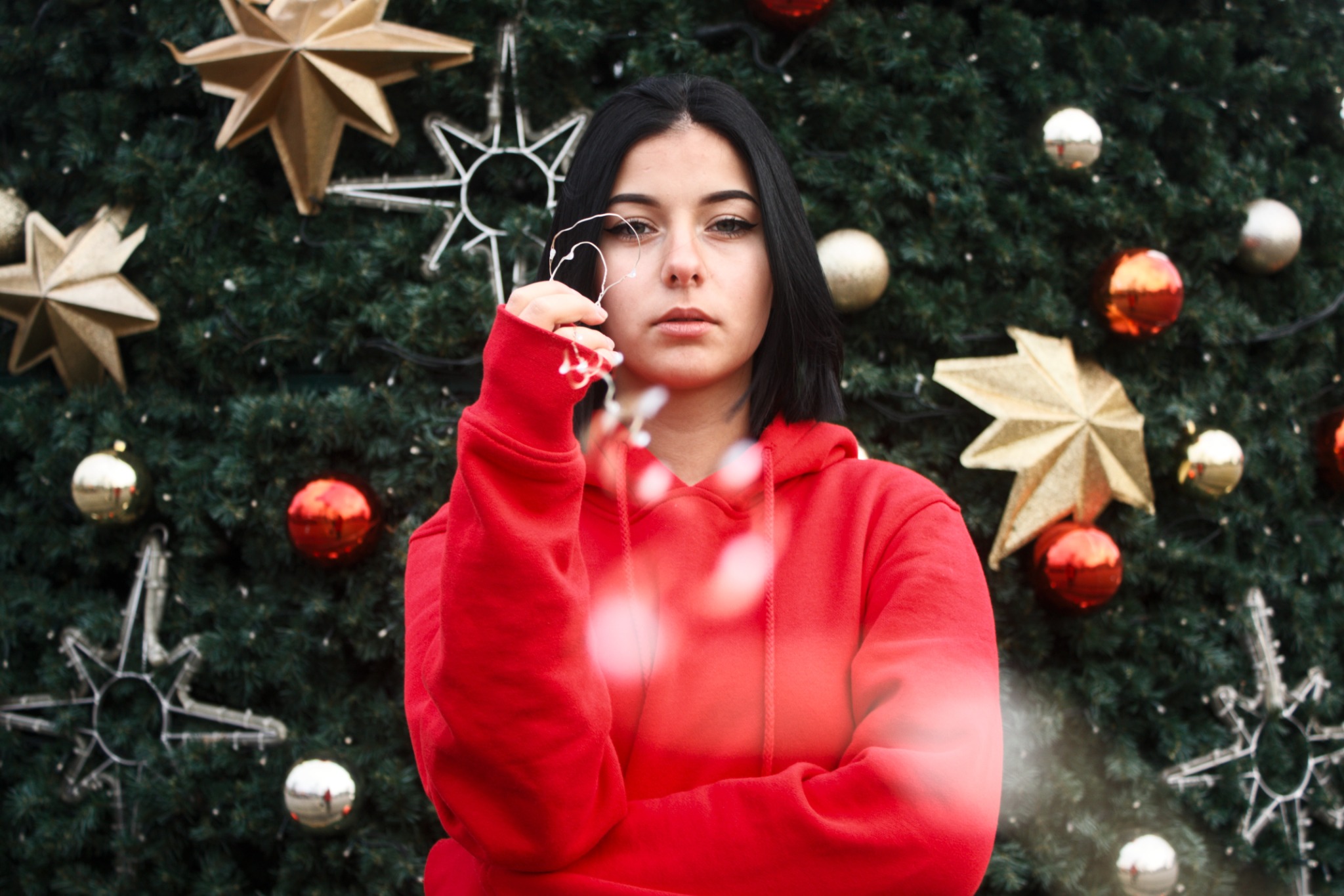 Młoda kobieta w czerwonej bluzie z kapturem pozuje przed choinką ozdobioną gwiazdami i bombkami w Dębicy, trzymając w dłoni światełka w kształcie serca.