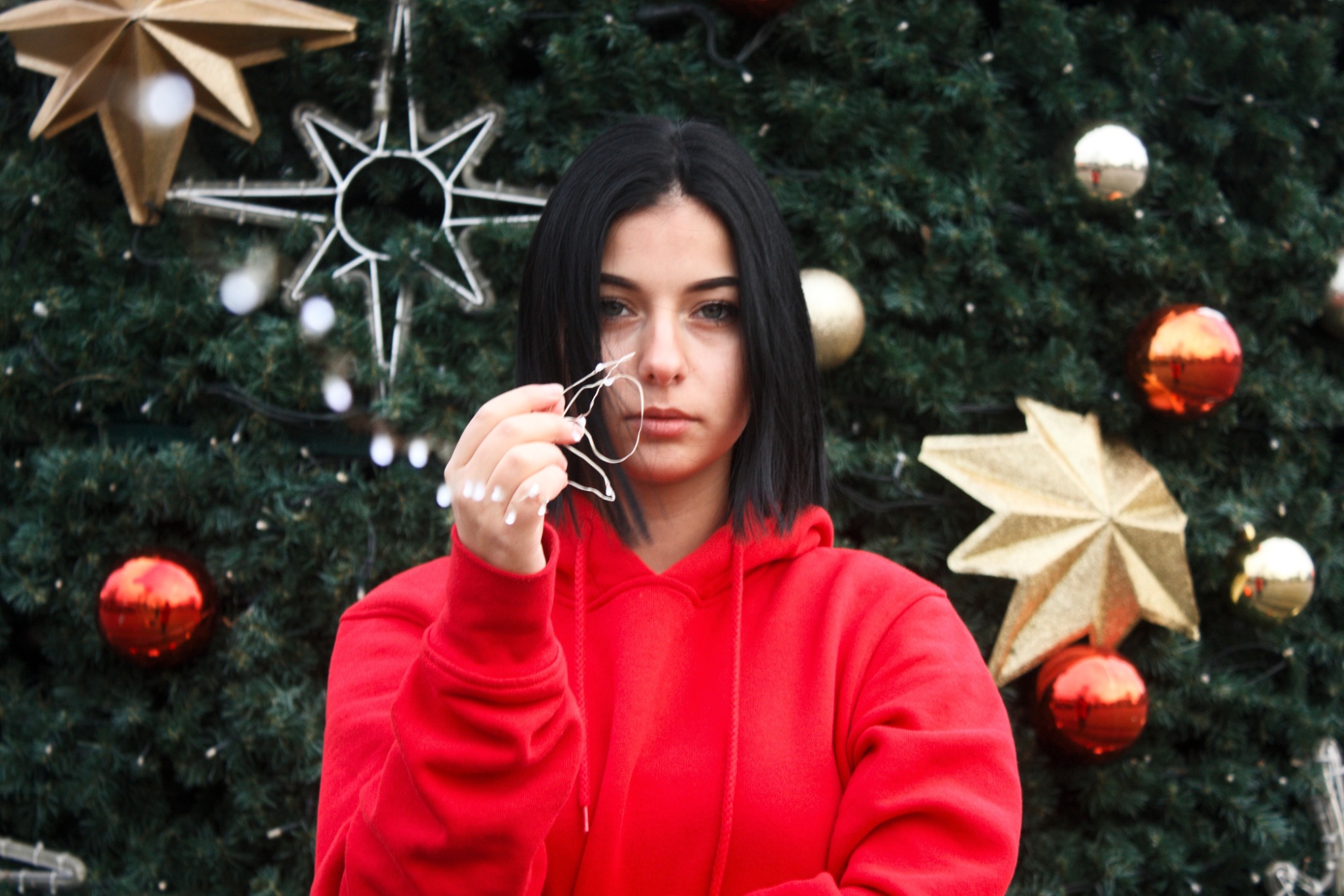 Kobieta w czerwonej bluzie z kapturem trzyma światełka choinkowe na tle udekorowanej choinki