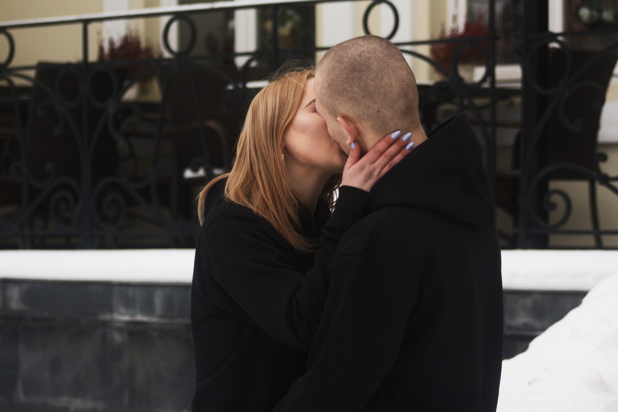 Zakochana para całuje się na tle budynku z ozdobną balustradą, kobieta z blond włosami i manicure obejmuje mężczyznę w czarnej bluzie z kapturem.
