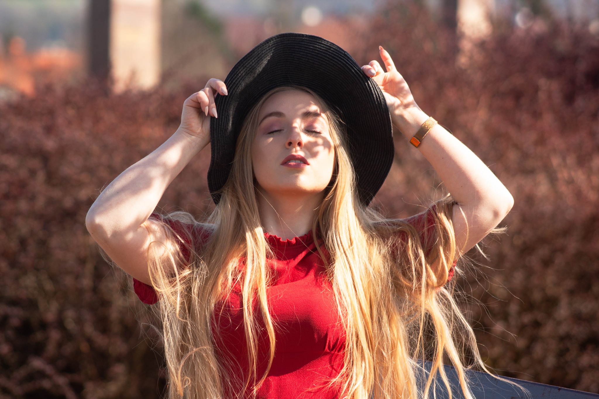Kobieta w czerwonej bluzce i czarnym kapeluszu z zamkniętymi oczami, dłonie uniesione ku górze, na twarzy cień, w tle rozmyte krzewy.