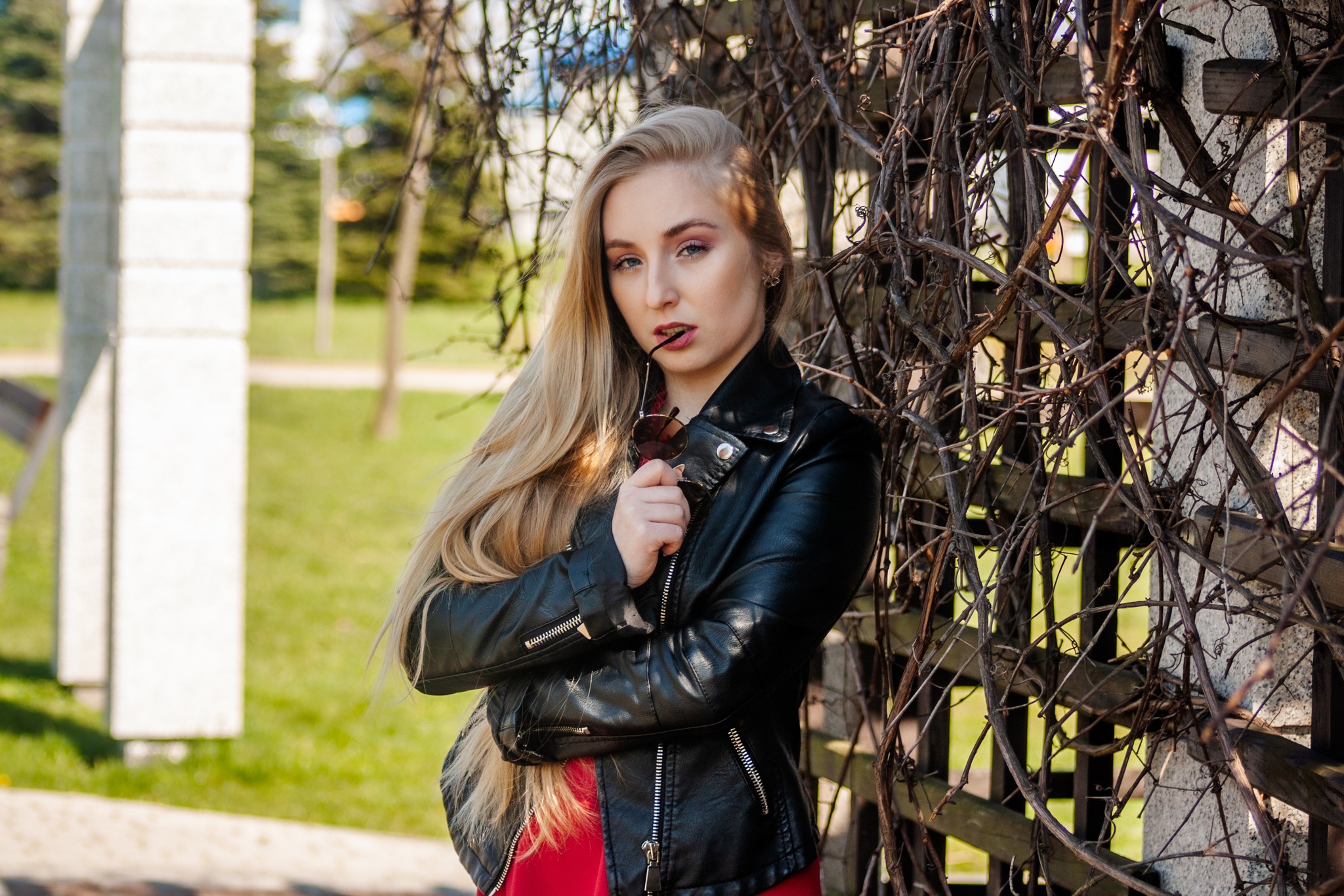 Portret blondwłosej kobiety w czarnej skórzanej kurtce, trzymającej okulary przeciwsłoneczne przy ustach, pozującej przy drewnianym płocie porośniętym suchymi pnączami na tle parku.