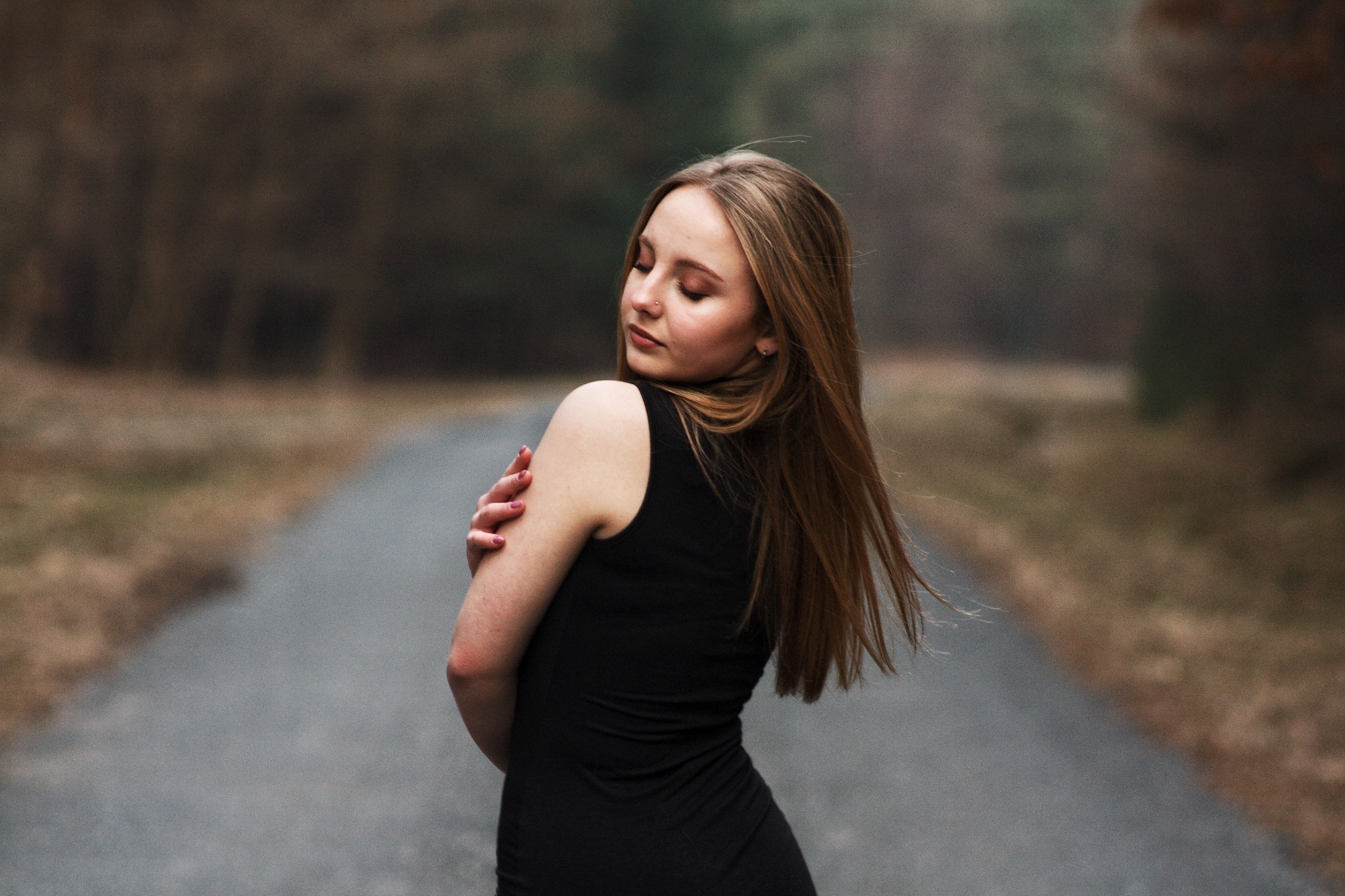 Kobieta w czarnej sukience pozuje na leśnej drodze, z rękami skrzyżowanymi na ramionach i głową odwróconą w bok.