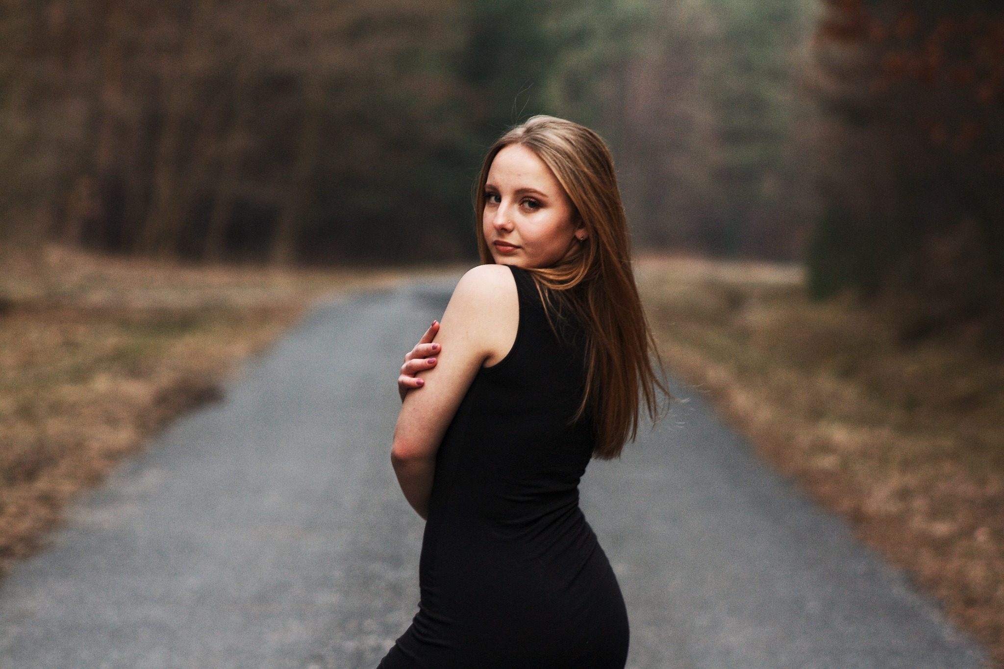 Portret kobiety w czarnej sukience, odwróconej przez ramię, na tle leśnej drogi w pochmurny dzień.