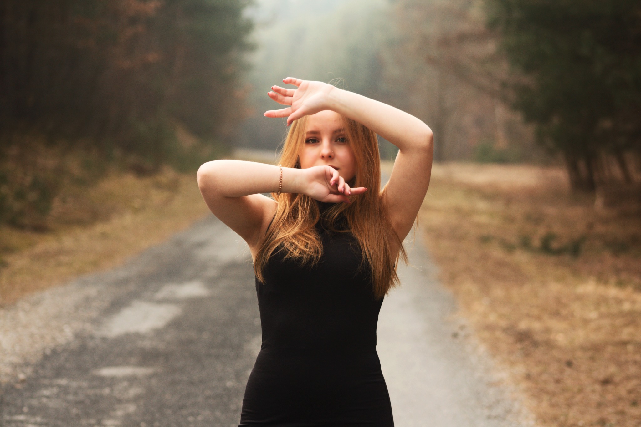 Młoda kobieta w czarnej sukience pozuje na leśnej drodze, dłońmi zasłania twarz, delikatne światło pada z góry.