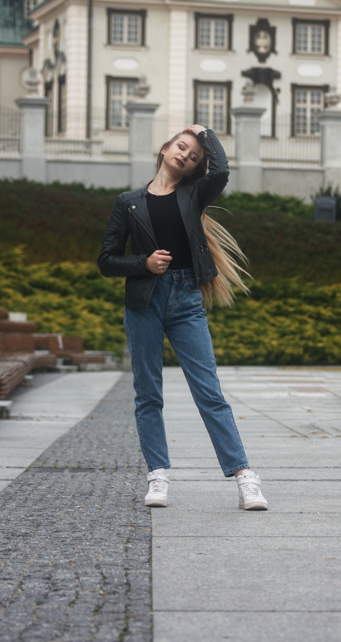 Młoda kobieta w czarnej skórzanej kurtce i jeansach pozuje na tle klasycystycznego budynku z zielenią, dłonią poprawia długie blond włosy.