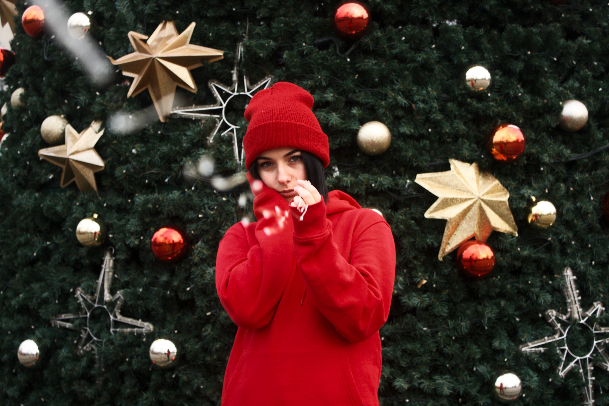 Młoda kobieta w czerwonej czapce i bluzie na tle choinki z ozdobami, trzymająca w dłoniach sztuczny śnieg.