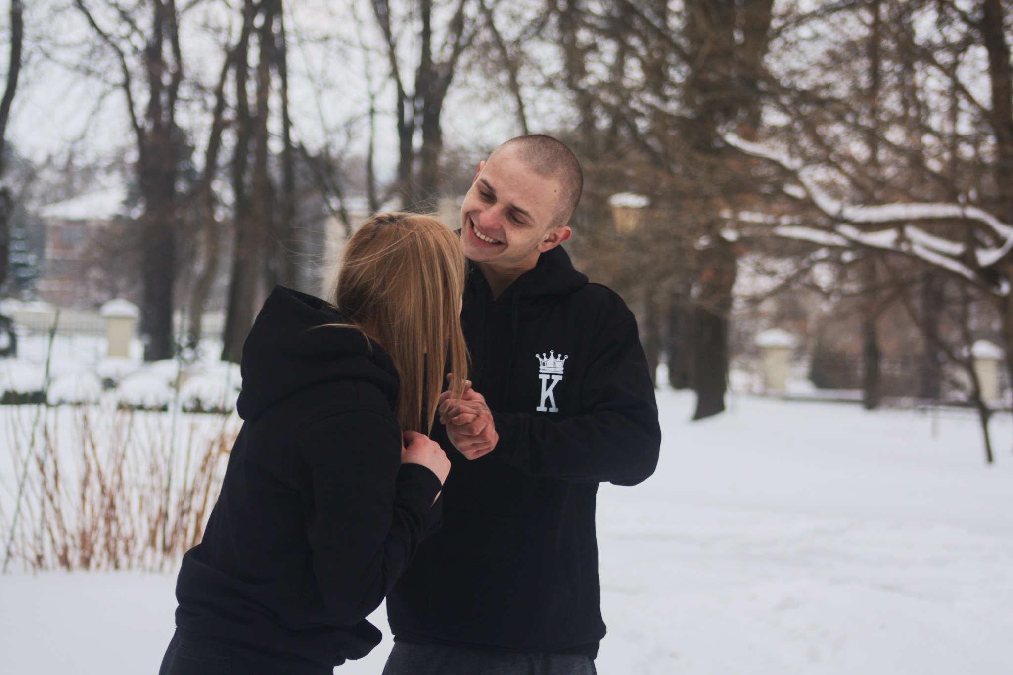 Uśmiechnięty mężczyzna w czarnej bluzie z kapturem, z logo korony i literą K, trzymający dłoń blondwłosej kobiety w zimowym parku pokrytym śniegiem.