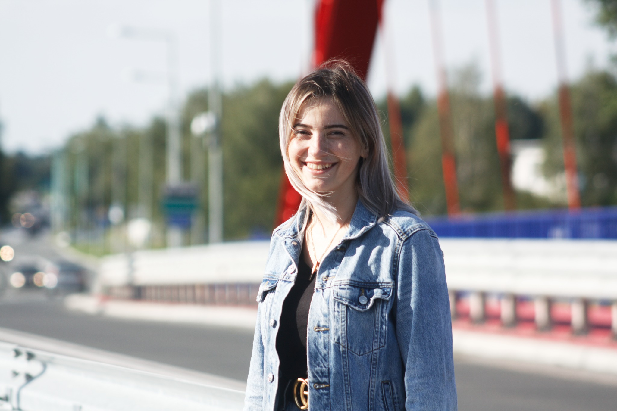Uśmiechnięta młoda kobieta w jeansowej kurtce, z platynowymi włosami, pozuje na moście z czerwonymi elementami konstrukcyjnymi w tle.