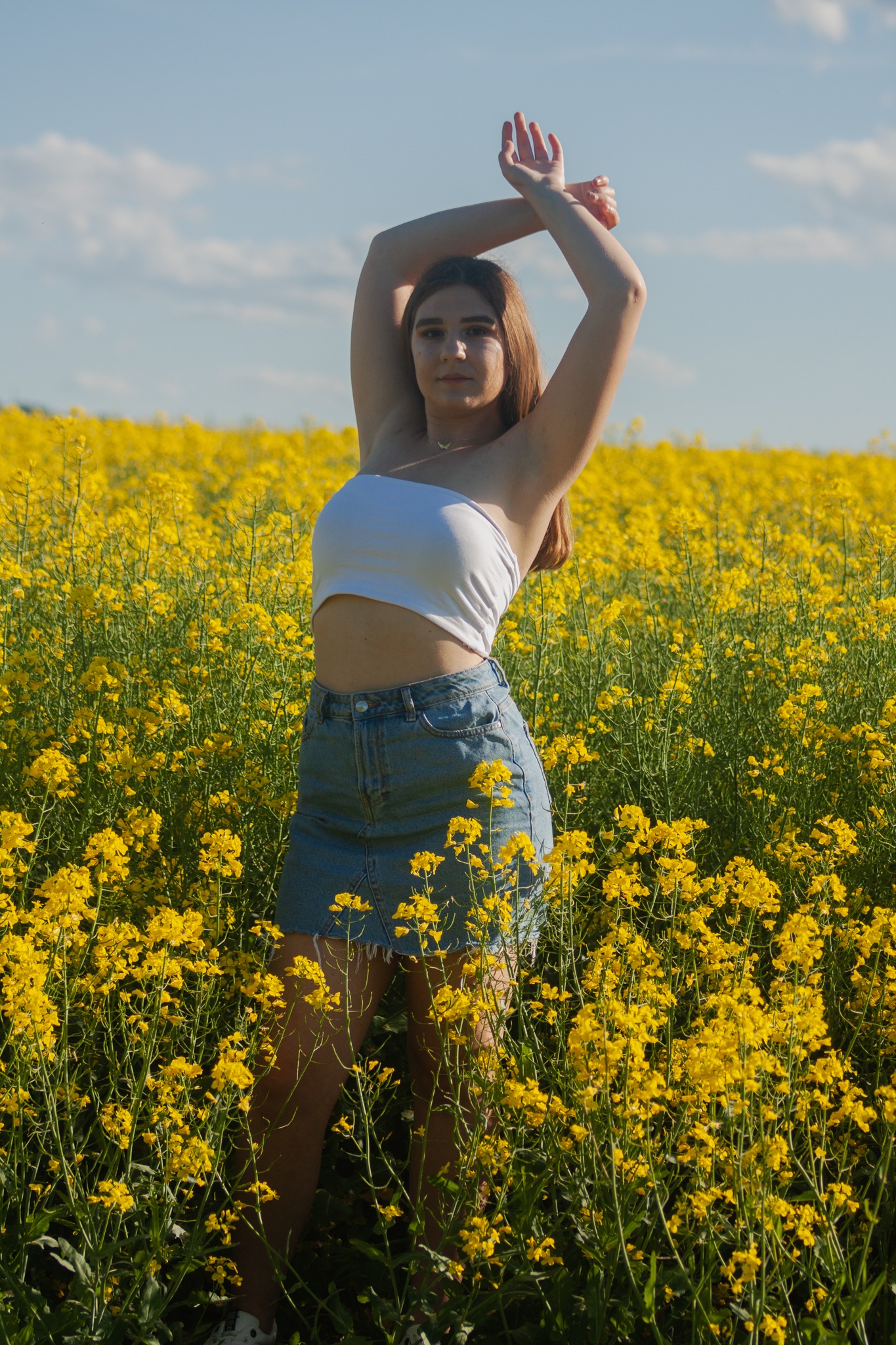 Młoda kobieta w białym topie i dżinsowej spódnicy pozuje z rękami uniesionymi nad głową na tle kwitnącego pola rzepaku w słoneczny dzień.