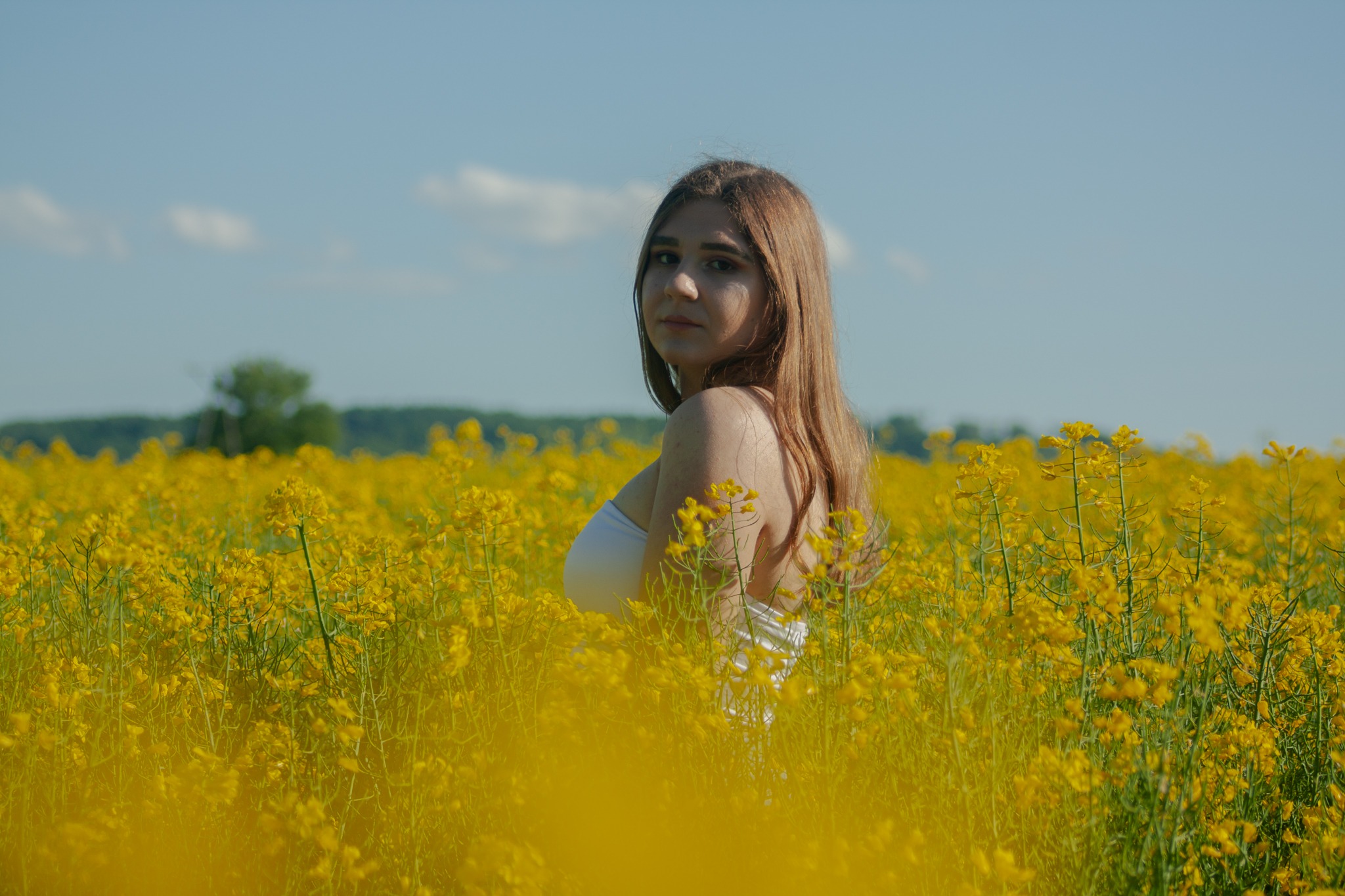 Kobieta w białej sukience spogląda przez ramię, stojąc pośród kwitnącego, żółtego pola rzepaku, z zalesionym horyzontem pod błękitnym niebem.