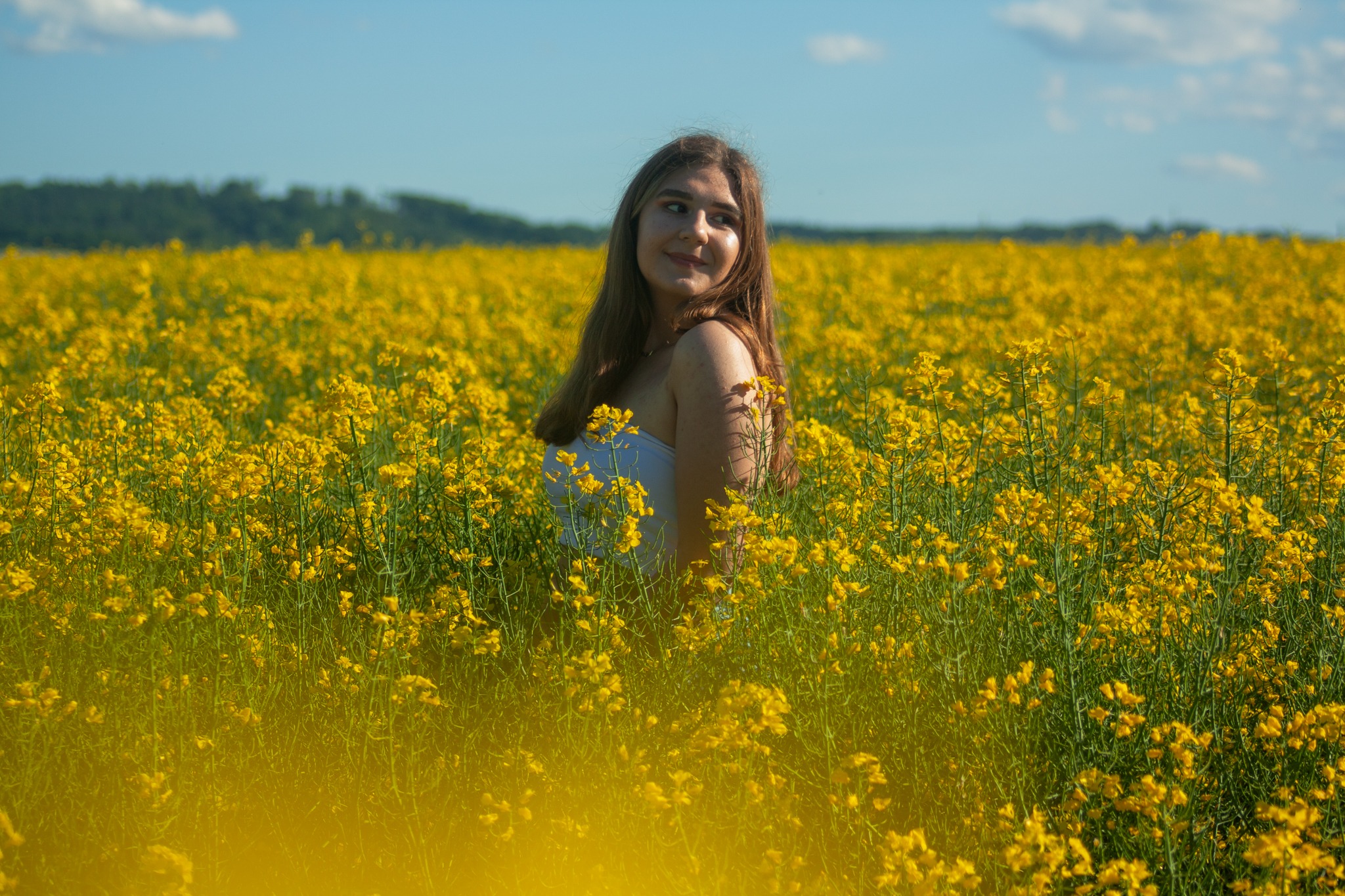 Portret uśmiechniętej kobiety w białej bluzce, stojącej na tle rozległego pola kwitnącego rzepaku w słoneczny dzień.