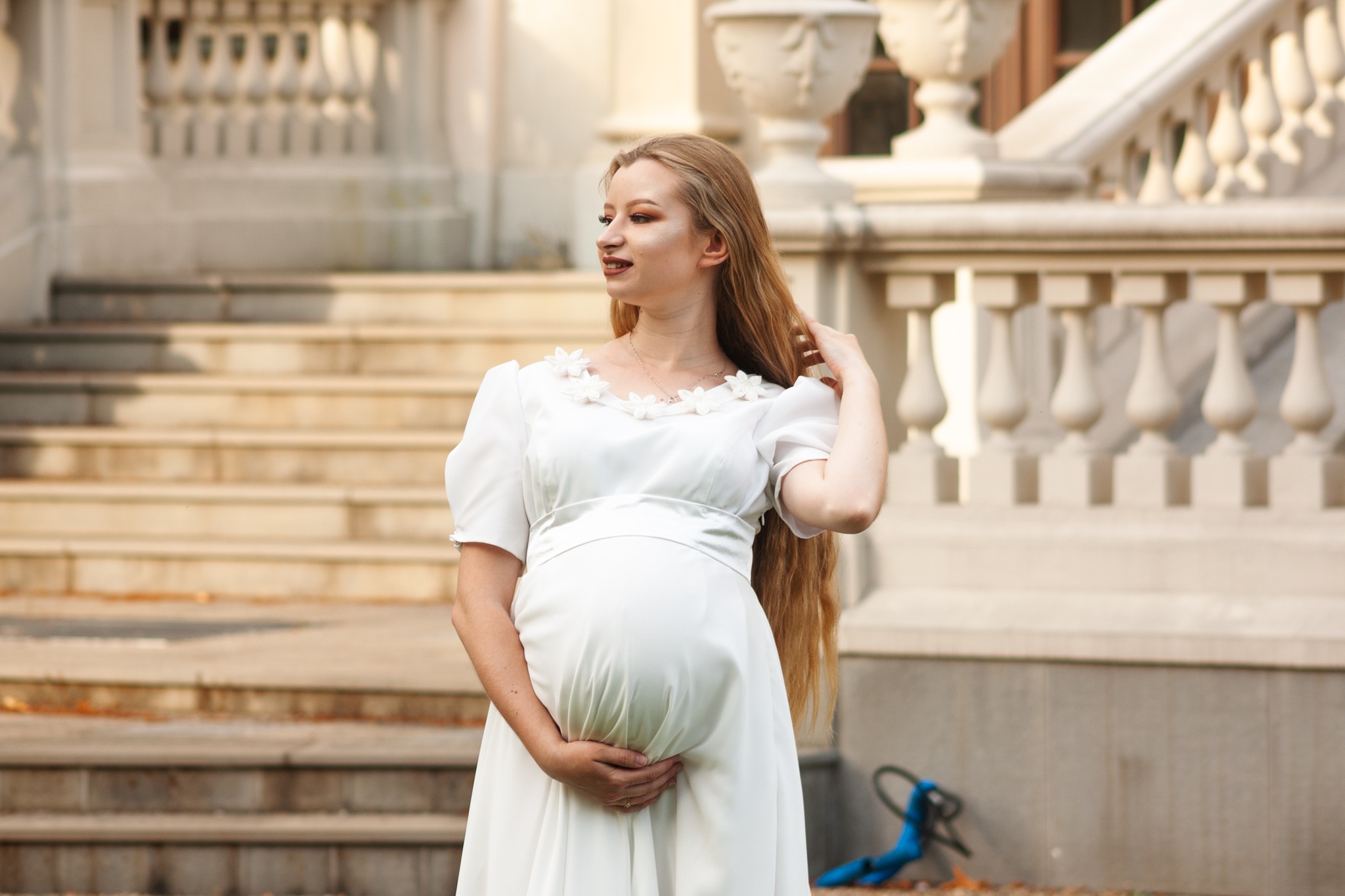 Kobieta w zaawansowanej ciąży, w białej sukni z kwiatami przy dekolcie, pozuje na tle klasycznej architektury z balustradami i schodami.