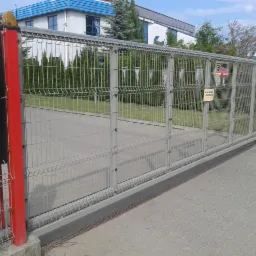 Przesuwna brama panelowa z czerwoną kolumną, widoczna tabliczka ostrzegawcza i budynek w tle. Brama wjazdowa z siatki, szara nawierzchnia.