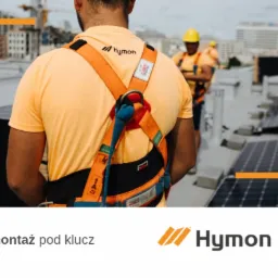 Instalator w uprzęży zabezpieczającej, widok z tyłu, na dachu budynku podczas montażu paneli słonecznych; w tle zabudowa miejska i drugi pracownik w żółtym kasku.