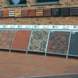 Ekspozycja różnych wzorów i kolorów pokryć dachowych na stojakach przed budynkiem z cegły klinkierowej, z dodatkowymi próbkami zamontowanymi na ścianie.