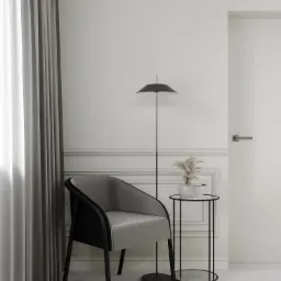 Minimalistyczny kącik do czytania: szary fotel z czarnymi nogami, czarna lampa podłogowa i okrągły stolik z wazonem z suszonymi trawami na tle białej ściany z dekoracyjnymi panelami i zasłoną.