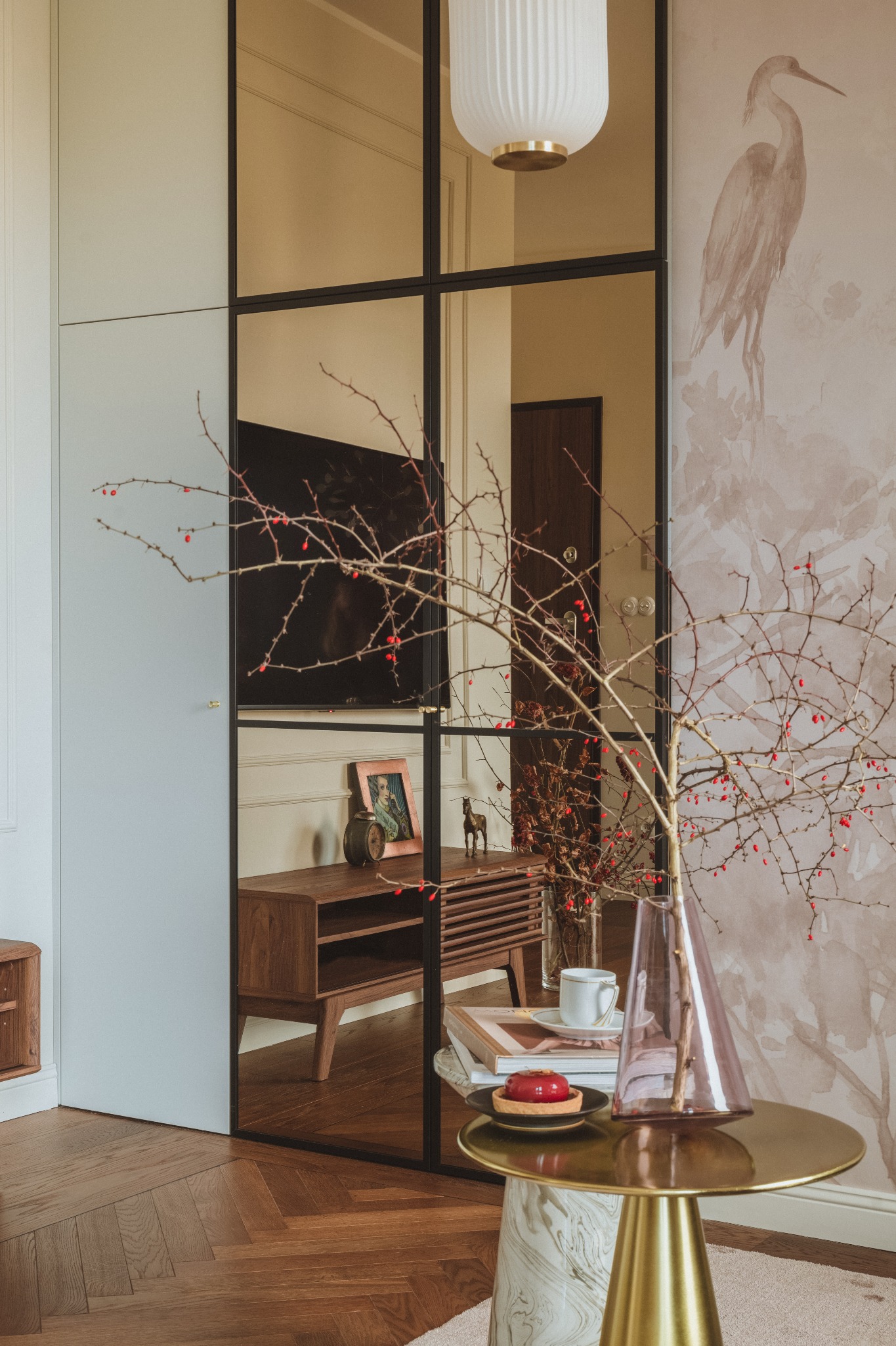 Eleganckie wnętrze z drewnianą komodą widoczną przez szklaną ścianę z czarnymi ramami, na pierwszym planie stolik z wazonem z gałązkami jarzębiny, książkami, filiżanką i deserem, w tle tapeta...