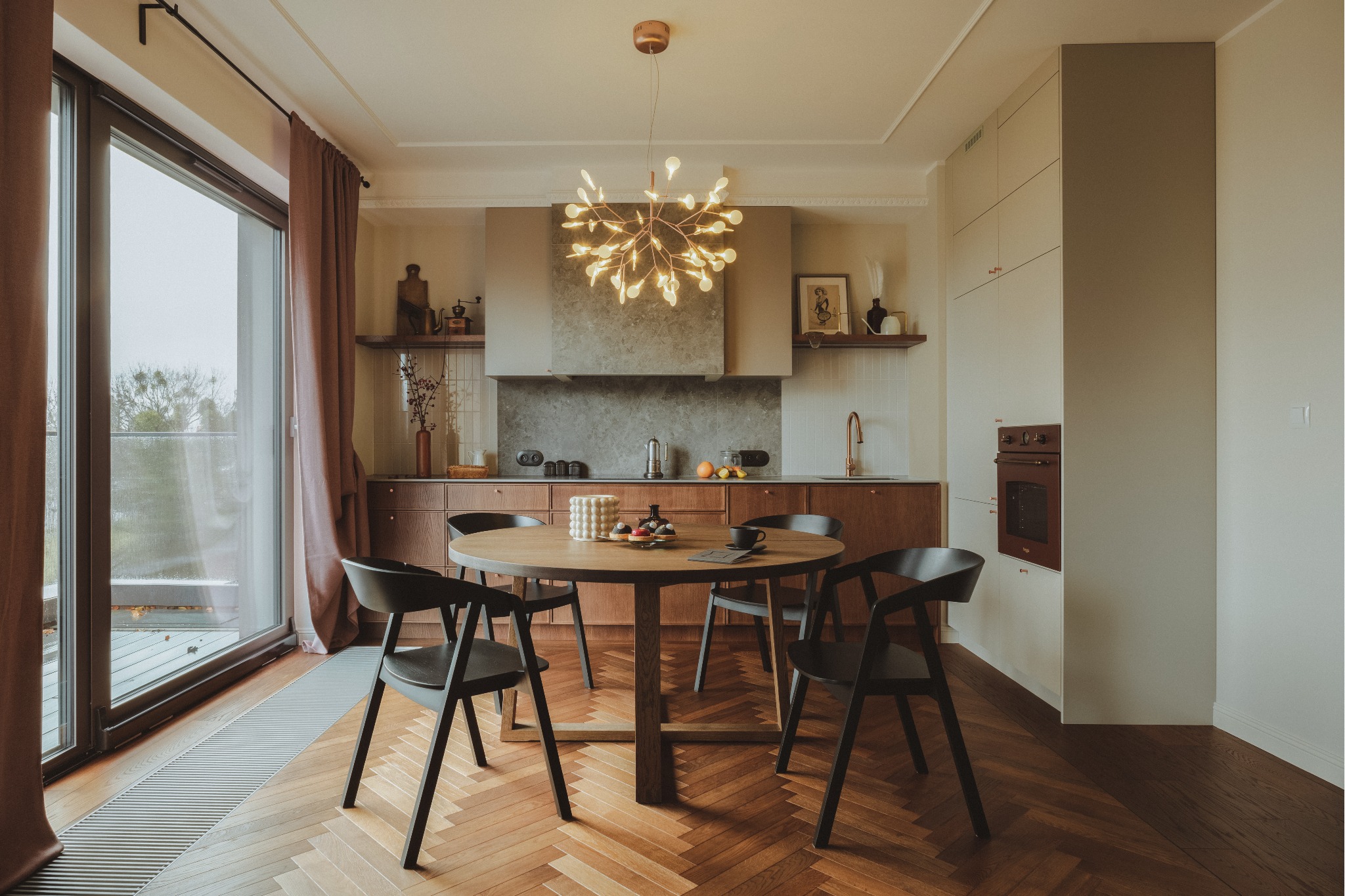 Jasna kuchnia z drewnianymi szafkami, okrągłym stołem z czarnymi krzesłami, parkietem w jodełkę i designerską lampą sufitową w kształcie rozgałęzionego drzewa.
