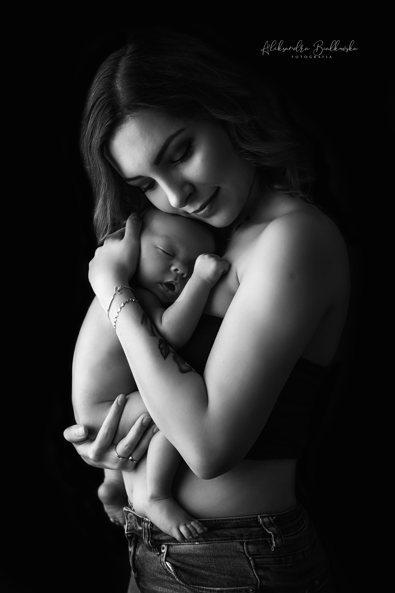 Czarno-białe zdjęcie matki czule obejmującej śpiące niemowlę, z widocznym tatuażem na ramieniu matki i jeansami z niskim stanem, na ciemnym tle.