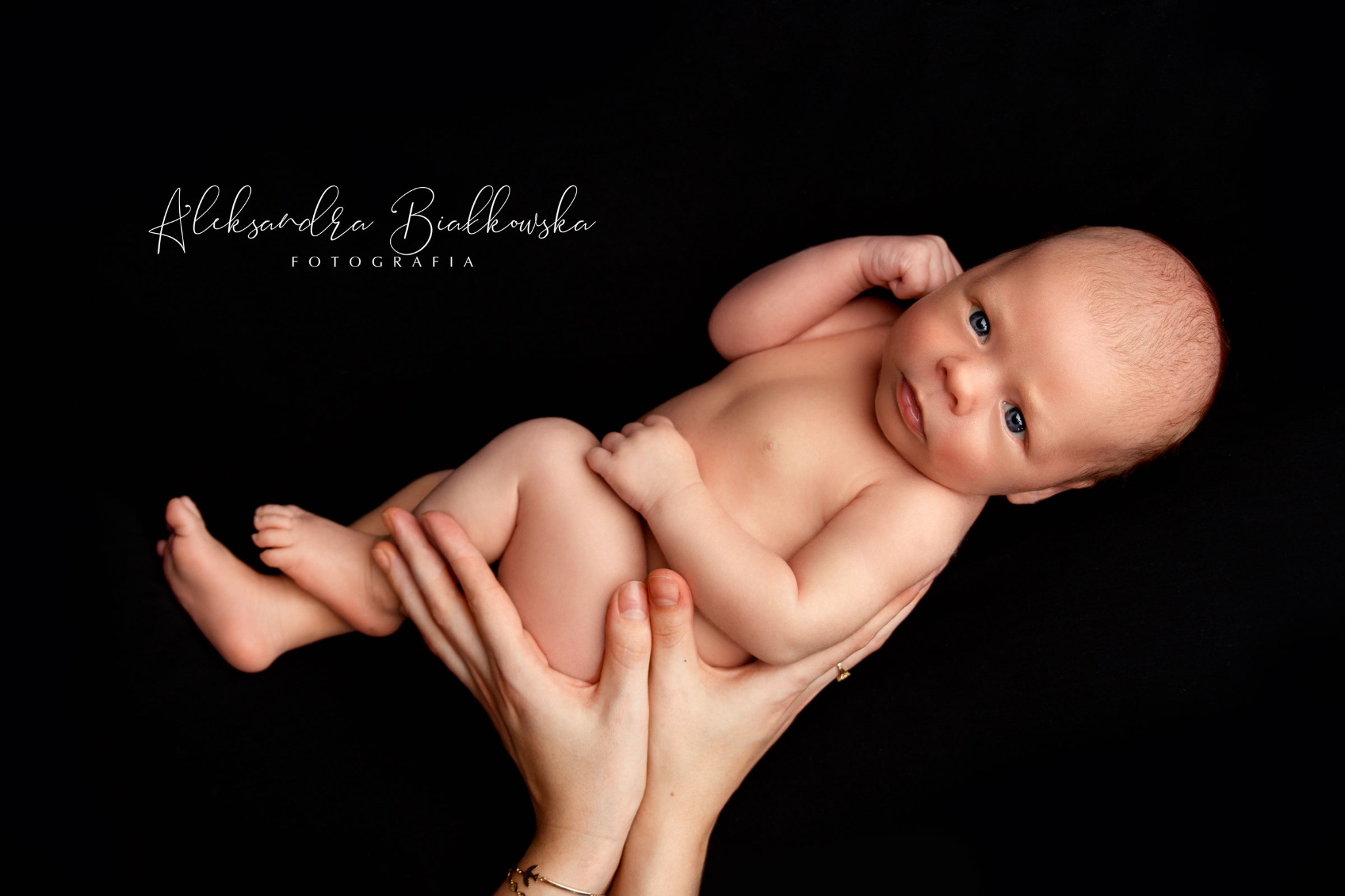 Noworodek z niebieskimi oczami, trzymany w dłoniach na czarnym tle, z widocznym logo fotografa.