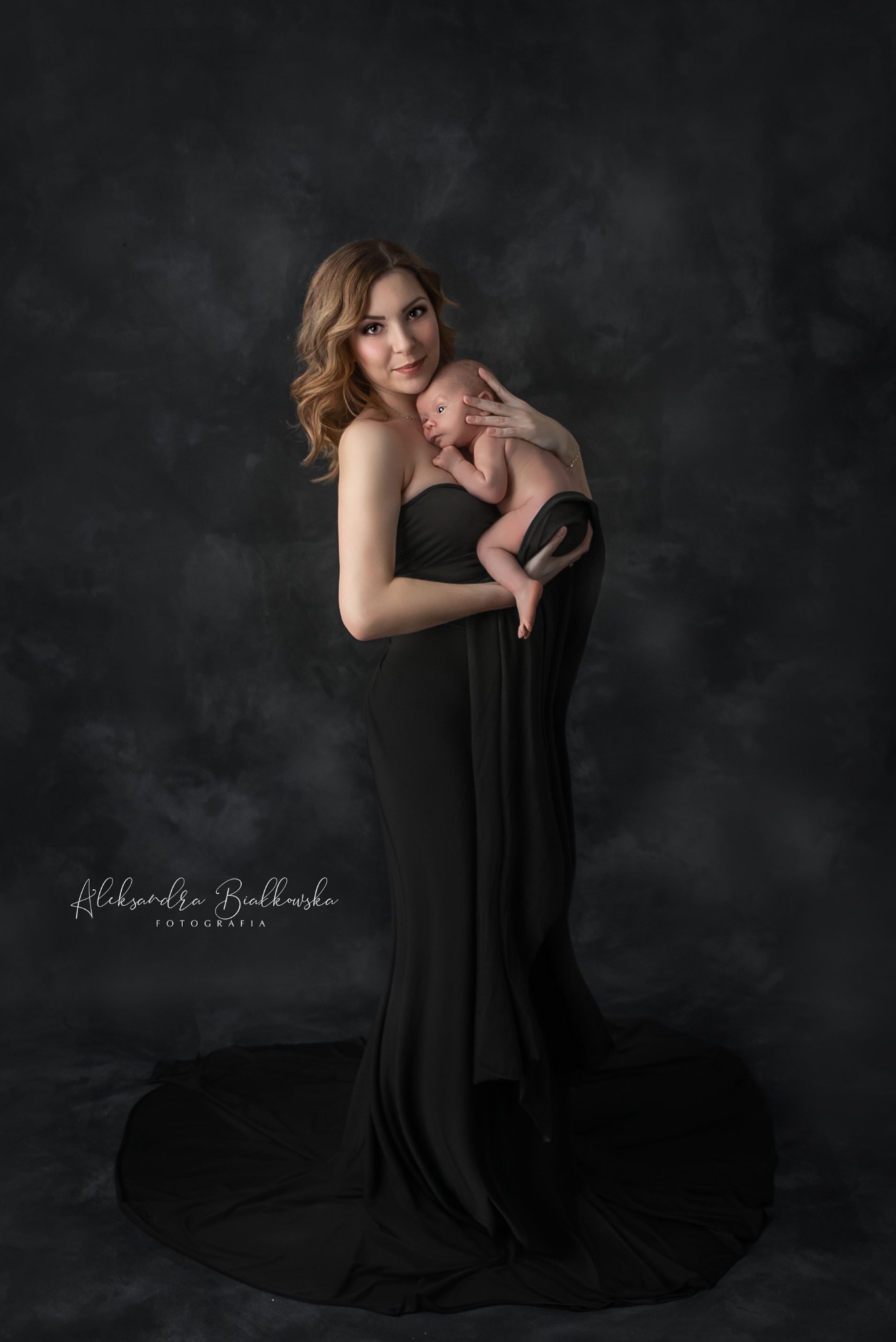 Elegancka kobieta w czarnej sukni trzyma noworodka na tle ciemnego, teksturowanego tła, ujęcie portretowe z delikatnym oświetleniem studyjnym.