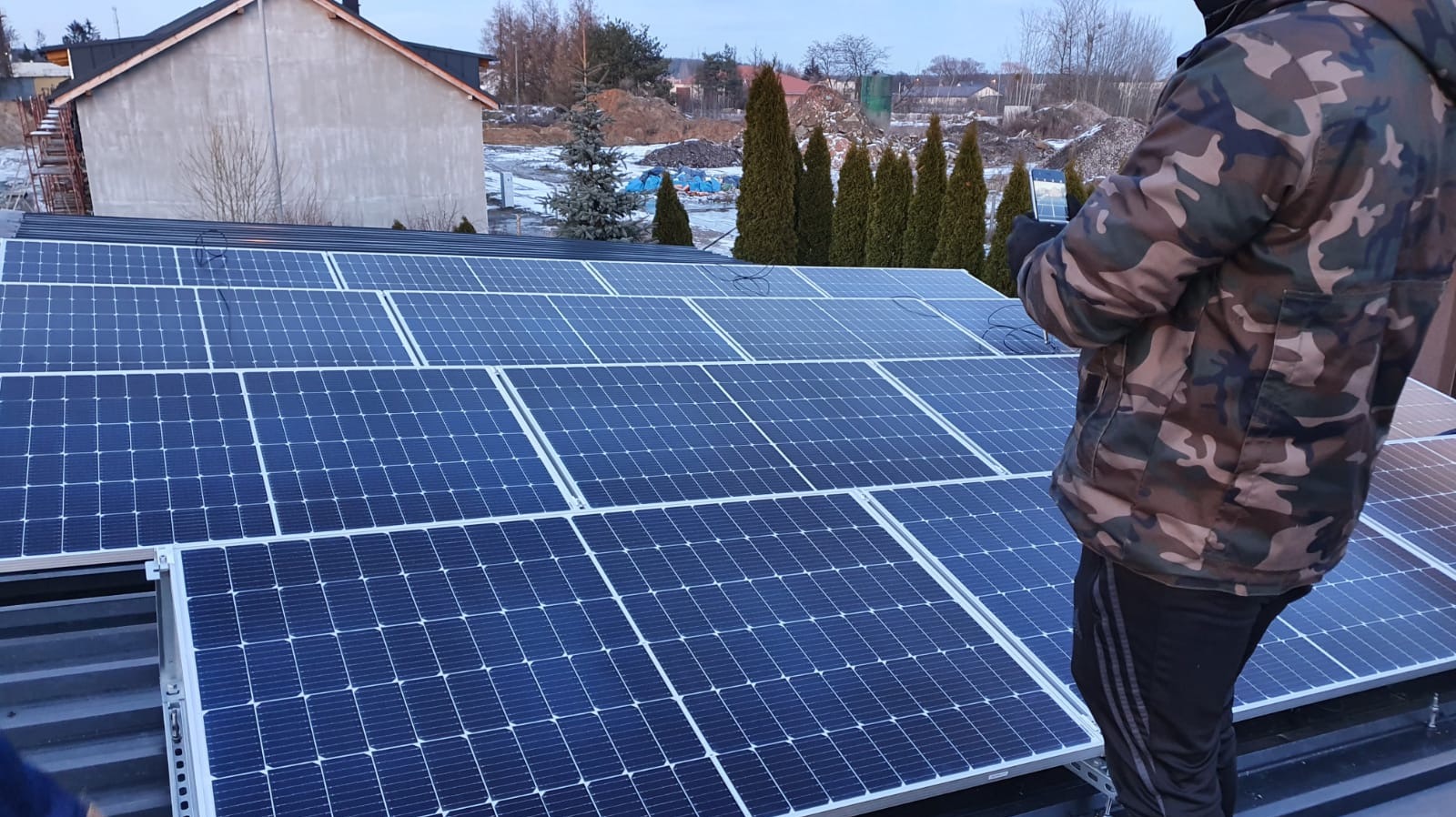 Montaż paneli słonecznych na dachu domu jednorodzinnego, widoczna osoba w kurtce moro podczas podłączania przewodów, w tle drzewa i budynki.