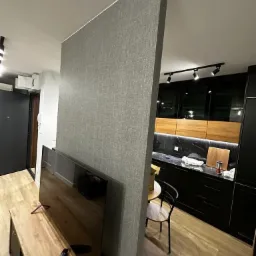 Szara ścianka działowa z wykończeniem tekstylnym w nowoczesnym wnętrzu, oddzielająca salon od kuchni z czarnymi szafkami i drewnianym blatem. Widoczny telewizor na drewnianej szafce RTV...