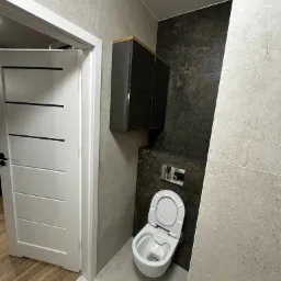 Nowoczesna łazienka z szarymi płytkami imitującymi kamień, wiszącą szafką w kolorze grafitowym z drewnianym blatem i toaletą podwieszaną z chromowanym przyciskiem spłukiwania.