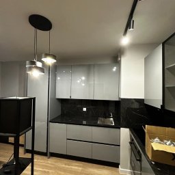 Bur-Bud - Nowoczesna kuchnia z szarymi, lakierowanymi frontami szafek, czarnym blatem imitującym marmur, oświetleniem szynowym i wiszącymi lampami nad wyspą, częściowo rozpakowana z kartonami na blacie.