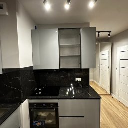 Bur-Bud - Nowoczesna kuchnia z szarymi szafkami, czarnym blatem imitującym marmur i drewnopodobną podłogą; oświetlenie punktowe sufitowe.