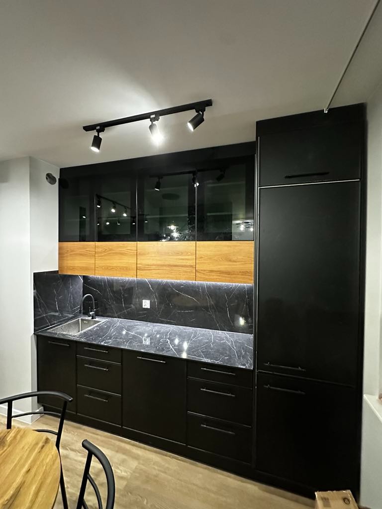 Nowoczesna kuchnia z czarnymi szafkami i blatem imitującym marmur, oświetlona listwą z reflektorami.