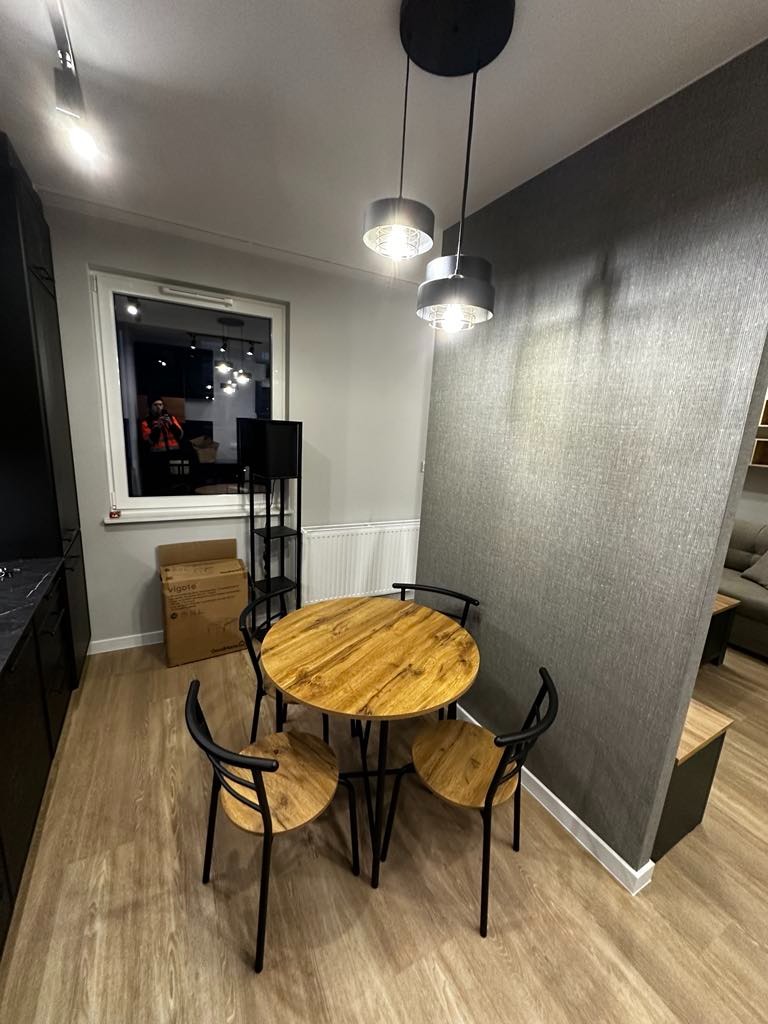 Jadalnia z okrągłym drewnianym stołem i czterema krzesłami na drewnianej podłodze; ściana wykończona szarą tapetą strukturalną; dwie nowoczesne lampy wiszące z metalowymi kloszami.