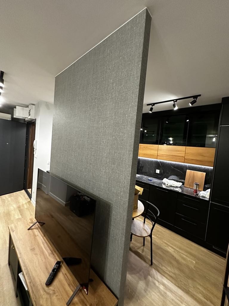 Szara ścianka działowa z wykończeniem tekstylnym w nowoczesnym wnętrzu, oddzielająca salon od kuchni z czarnymi szafkami i drewnianym blatem. Widoczny telewizor na drewnianej szafce RTV...