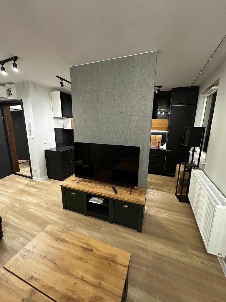 Nowoczesny salon z drewnianą podłogą imitującą panele, ciemnozieloną szafką RTV i szarą ścianką działową, minimalistyczne oświetlenie punktowe.