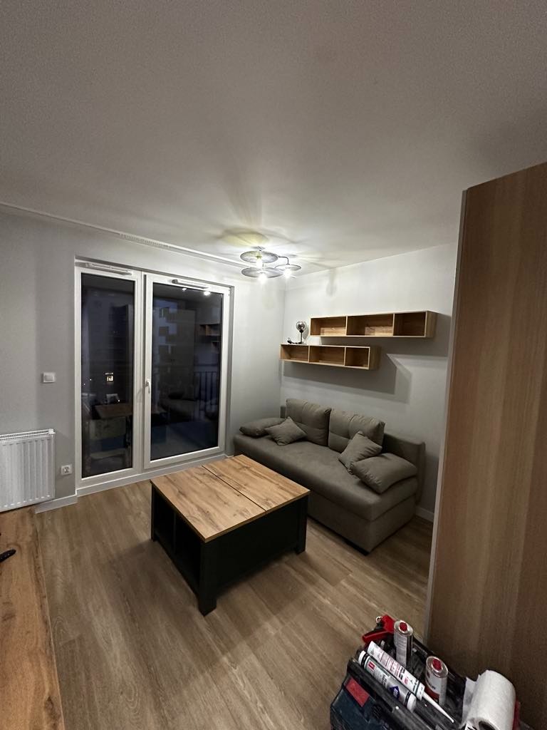 Salon po remoncie z panelami podłogowymi w odcieniach drewna, szarą sofą i czarno-drewnianym stolikiem kawowym, widoczne narzędzia i materiały budowlane w rogu pomieszczenia.