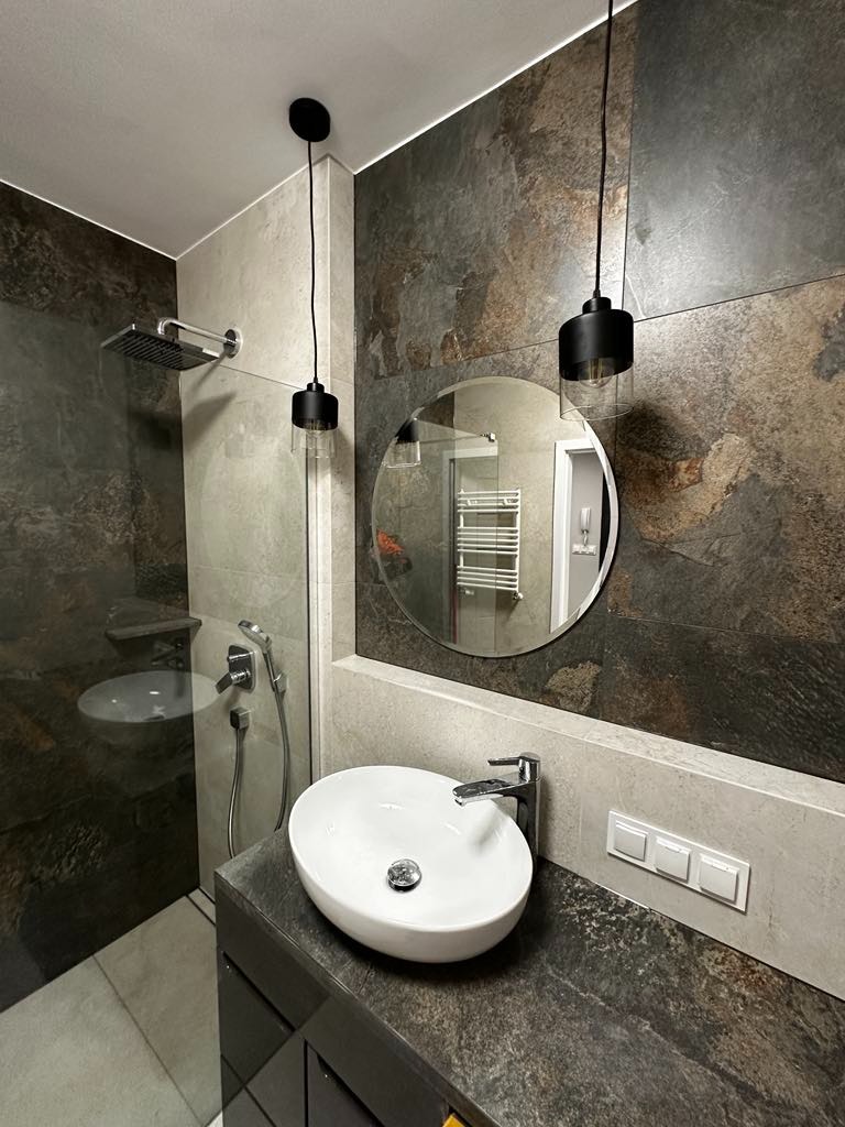 Nowoczesna łazienka z kabiną prysznicową typu walk-in, umywalką nablatową o owalnym kształcie, okrągłym lustrem i dwoma wiszącymi lampami z czarnymi kloszami i szklanymi elementami, wykończona...