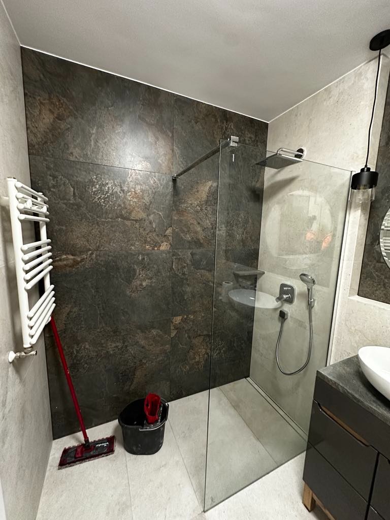 Nowoczesna łazienka z kabiną prysznicową typu walk-in, wykończona płytkami imitującymi kamień w odcieniach szarości i brązu, z widocznym mopem i wiadrem w rogu.