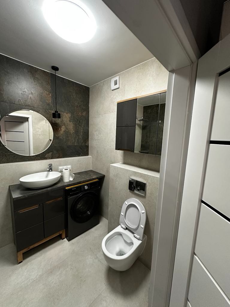 Nowoczesna łazienka z szarą armaturą, podwieszana toaleta z otwartą deską, pralka obok umywalki nablatowej i lustro okrągłe z czarną lampą wiszącą.