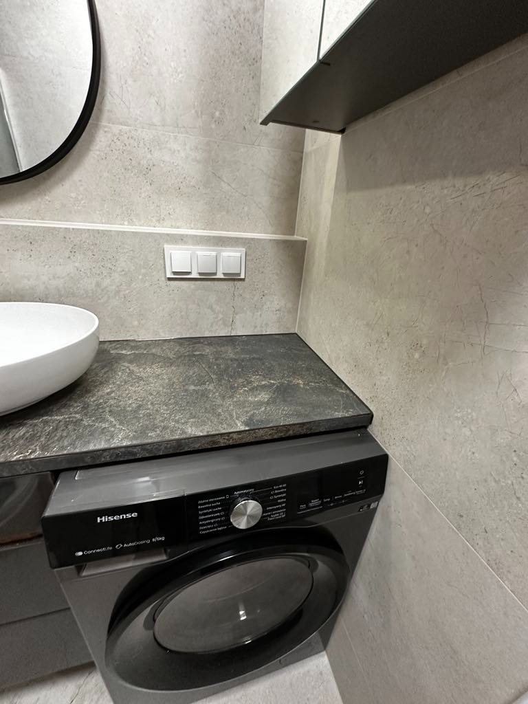 Szara łazienka z pralką Hisense pod blatem z ciemnego kamienia, umywalką nablatową i płytkami imitującymi kamień.