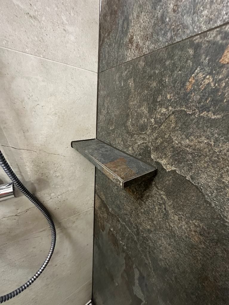 Półka z ciemnego kamienia w kabinie prysznicowej z szarymi płytkami, widoczny wąż prysznicowy.