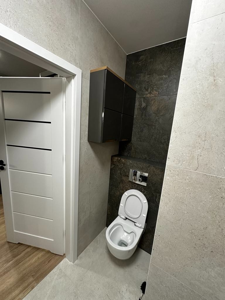 Nowoczesna łazienka z szarymi płytkami imitującymi kamień, wiszącą szafką w kolorze grafitowym z drewnianym blatem i toaletą podwieszaną z chromowanym przyciskiem spłukiwania.