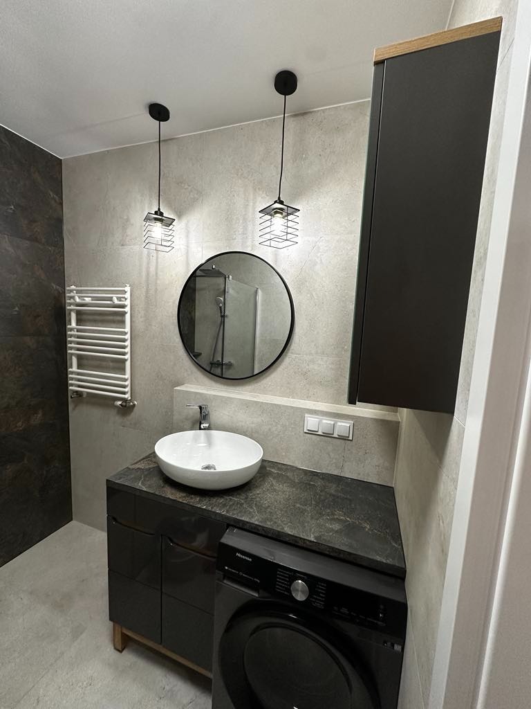 Nowoczesna łazienka w Żerkowie z okrągłym lustrem, wiszącą umywalką, ciemnymi szafkami i pralką, oświetlona dwoma designerskimi lampami wiszącymi w metalowych klatkach.